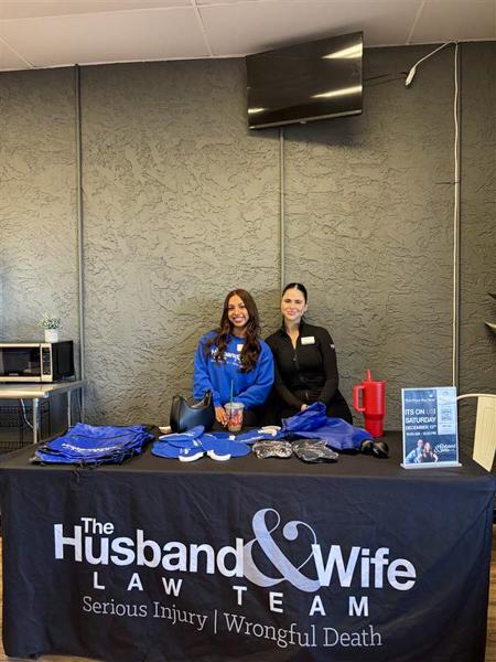 Two women stand behind a table with promotional materials and a banner reading "The Husband & Wife Law Team" in an office setting.