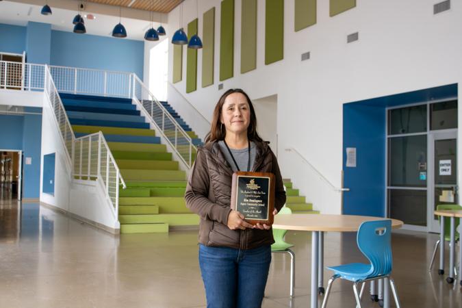 Woman standing in a bright school lobby holding an award plaque, with colorful tiered seating, stairs and blue chairs in the background.