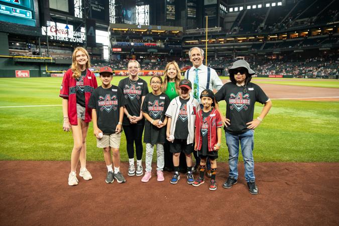 A group of children and adults stand on a baseball field, smiling. They wear team-themed clothing, with stadium seating visible in the background.