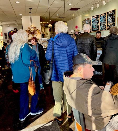 People gathered inside a cozy Tribes Coffeehouse, some seated and others standing in line near the counter with a menu board above.