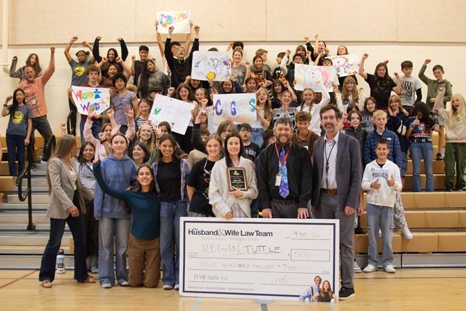 In the gymnasium, students and teachers gather with signs and a large check, with Ms. Tuttle prominently positioned at the front holding a plaque.