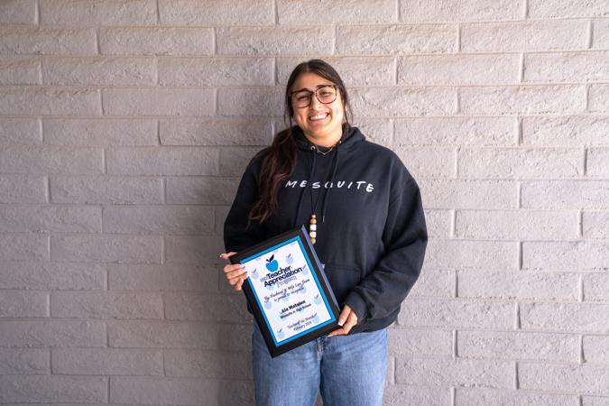 Ms. Matalon stands against a light brick wall at Mesquite Junior High, smiling as she holds her framed teacher appreciation certificate, wearing glasses and a dark hoodie.